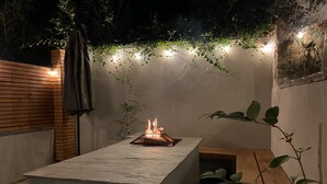 A rectangular outdoor seating area with a built-in fire feature, surrounded by plants and a wall with string lights. - Clovelly House: Relaxed Family Coastal Home (Berwick-upon-Tweed)