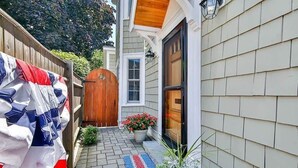A narrow outdoor pathway leading to a wooden door, with a flag and a potted plant on the left.
