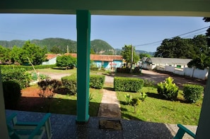 A view from a balcony with a green railing, overlooking a courtyard with neatly trimmed bushes, a pathway, and a building with a red-tiled roof. - Villa Gilda (Viñales)