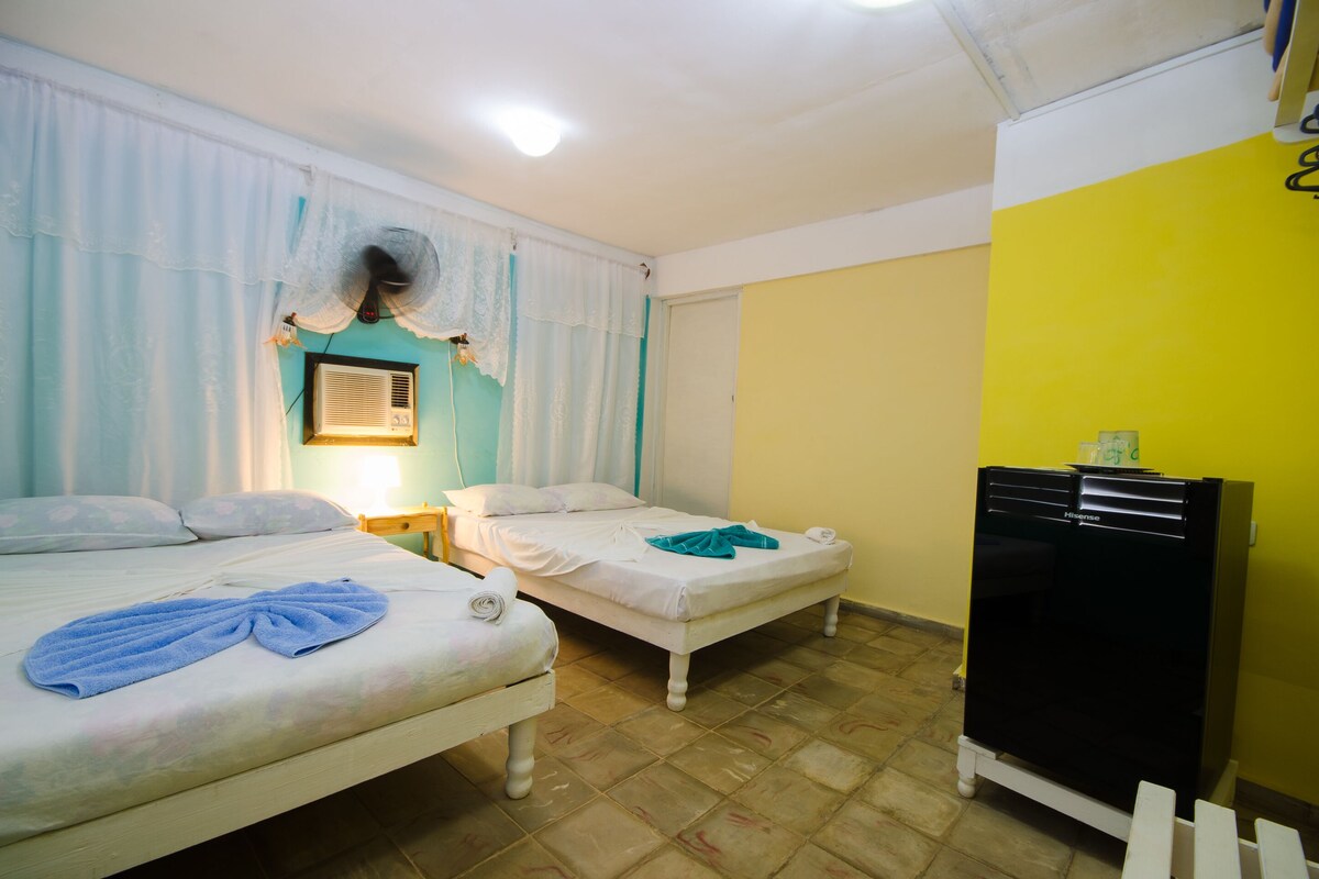 A room with two beds, a ceiling fan, a heater, and a refrigerator.