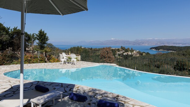 A swimming pool with lounge chairs and an umbrella, surrounded by stone tiles and a view of the sea and mountains. - Villa Anemelia by Rodostamo Collection (Kassiopi)