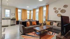 Ein modernes Wohnzimmer mit einer Couch, einem Couchtisch und einem Fernseher. Vorhanden sind Vorhänge, ein Teppich und eine Küche im Hintergrund.