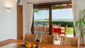 Un coin repas avec une table dressée pour deux personnes, une chaise en bois et une vue sur la mer par une porte vitrée.