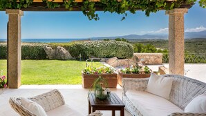 Un espace extérieur aménagé, abrité et doté de mobilier en osier, de plantes en pot, et offrant une vue sur la mer et les montagnes.