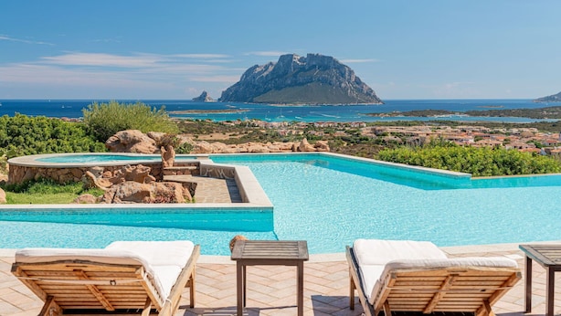 A pool area with lounge chairs, a stone structure, and a view of the sea and a distant island.