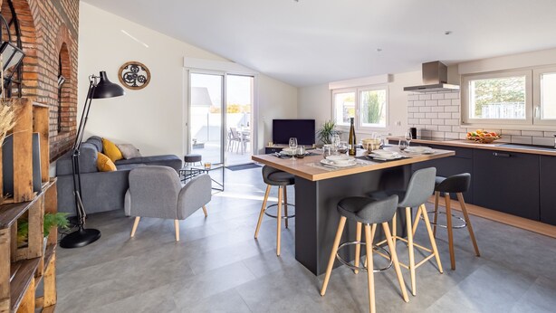 A modern kitchen with a central island, a dining area with a sofa and chairs, and a brick wall. - L'Atelier de notre Grand-Père - Jacuzzi (Algolsheim)