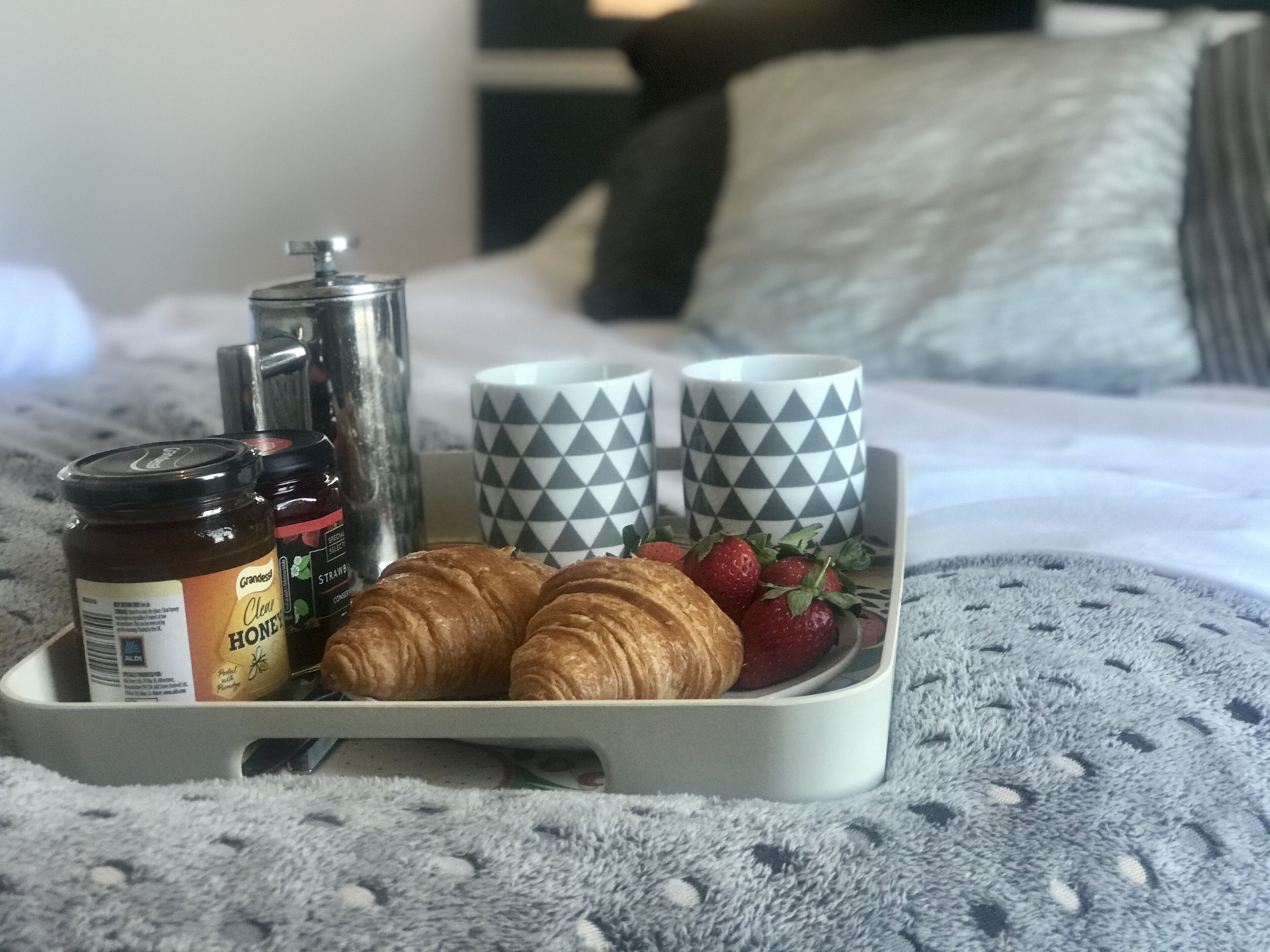 Un plateau de petit-déjeuner avec des croissants, des fraises, du miel et du café, posé sur un lit avec des oreillers et une couverture.