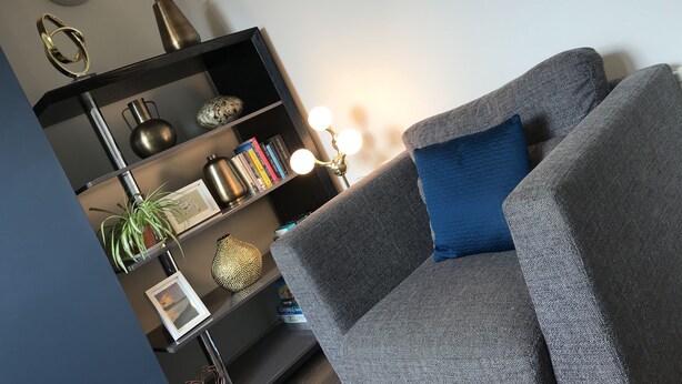 Un salon moderne avec un canapé gris, une bibliothèque, des objets de décoration et un luminaire fixé au mur.