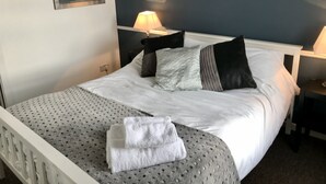 Un lit bien fait, avec du linge de lit blanc, un plaid à motifs gris et une serviette pliée. On y trouve deux lampes de chevet, une horloge murale et un tableau encadré au-dessus du lit.