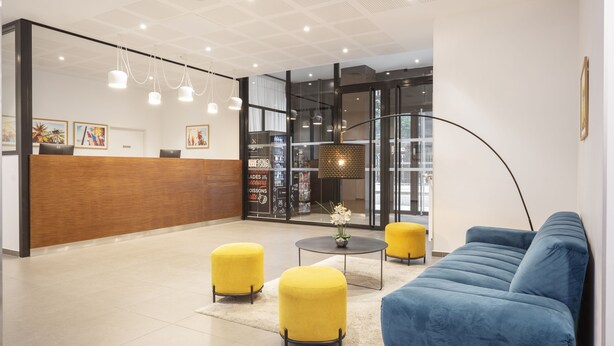 A modern reception area with a wooden reception desk, yellow ottomans, a blue sofa, and a large floor lamp. - Séjours & Affaires Paris Bagnolet (Bagnolet)