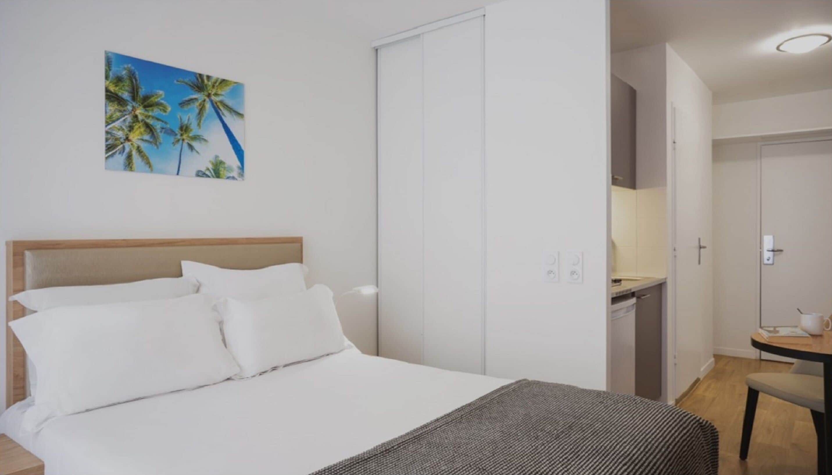 A bedroom with a bed, a nightstand, a painting of palm trees, a closet, and a kitchenette area.