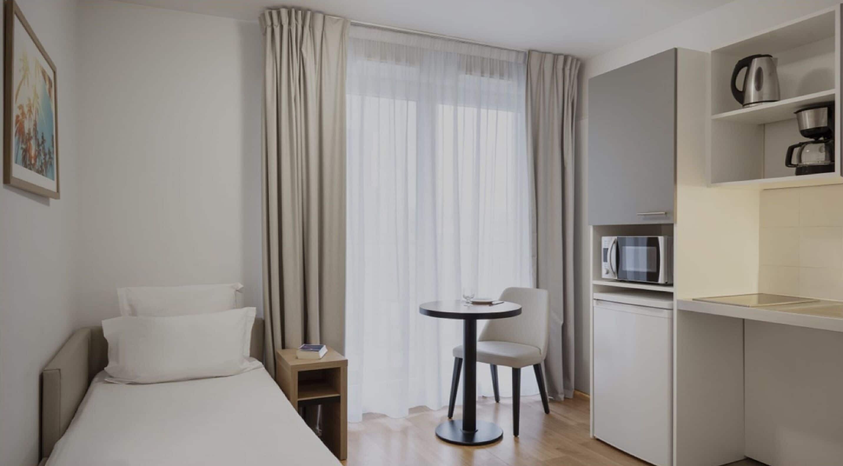 A hotel room with a bed, a small table, a chair, a kitchenette with a microwave, and a window with curtains.