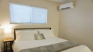 A neatly made bed with a gray throw blanket, a bedside lamp, and a window with blinds.