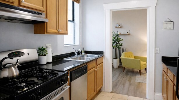 A modern kitchen with wooden cabinets, a stove, and a sink. There is a yellow chair in the adjacent living area.