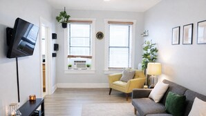 A living room with a flat-screen TV, two windows with blinds, a sofa, a chair, a lamp, and a small table with decorative items.