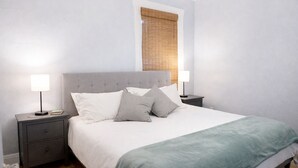 A neatly made bed with a grey headboard, two bedside tables with lamps, and a window with blinds. - Downtown Gem • King Bed • Fast WIFI (Salt Lake City)