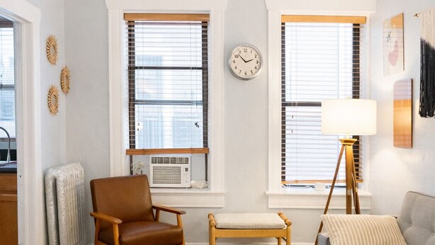 A living room with a sofa, armchair, and a small table. There are two windows with blinds, a radiator, and a clock on the wall. - Downtown Gem • King Bed • Fast WIFI (Salt Lake City)