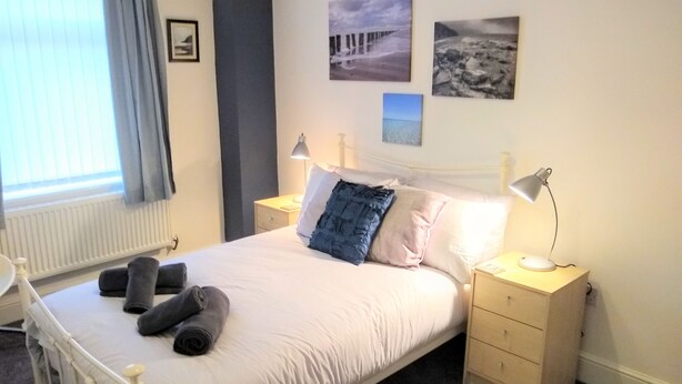 A neatly made bed with pillows and rolled towels, a bedside lamp, and a small bedside table with a lamp. - Modern Comfy 2-bedroom Flat in St Helens (St Helens)