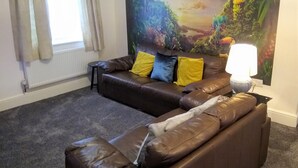 A living room with a brown leather sofa, two yellow cushions, a small side table, and a large painting of a forest scene. - Modern Comfy 2-bedroom Flat in St Helens (St Helens)