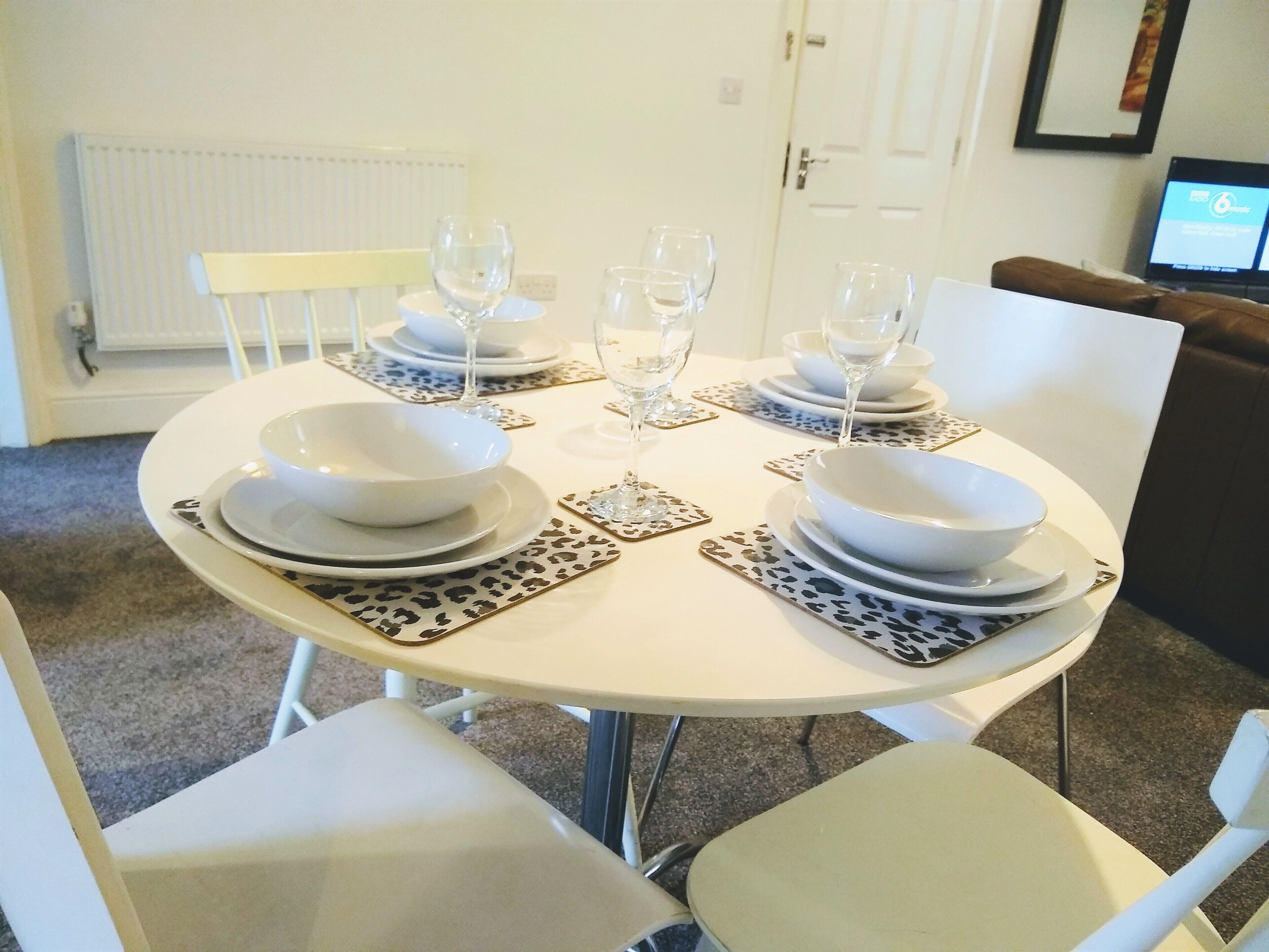 Una mesa de comedor dispuesta con platos, tazones y vasos blancos, rodeada de sillas con manteles individuales de estampado de leopardo.