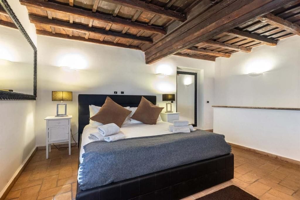 A bedroom with a large bed, wooden ceiling beams, a nightstand, and wall-mounted lights.