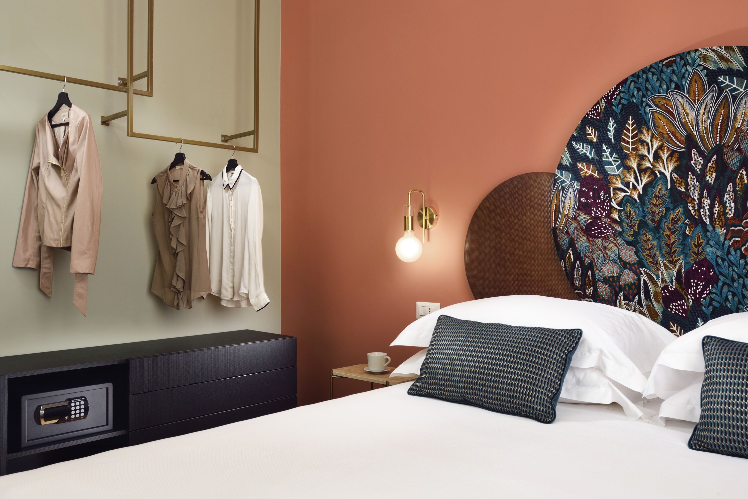 Habitación de hotel con cama, mesita de noche con una taza, perchero en la pared y una caja de seguridad.