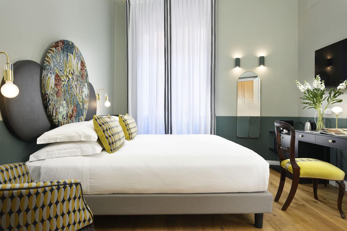 A bedroom with a bed, bedside lamps, a desk, a chair, a mirror, and a decorative wall piece.