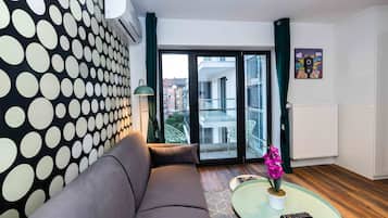 Ein modernes Wohnzimmer mit einem grauen Sofa, einem Glastisch mit einem Topf-Orchideenstrauß und Blick auf die Stadt durch große Glasfenster.
