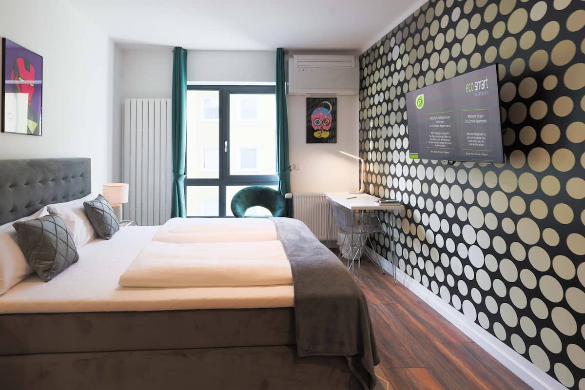 Ein modernes Schlafzimmer mit Bett, Schreibtisch und Stuhl. Ein Fenster mit Vorhängen, eine Heizung und eine Wandklimaanlage sind ebenfalls vorhanden.