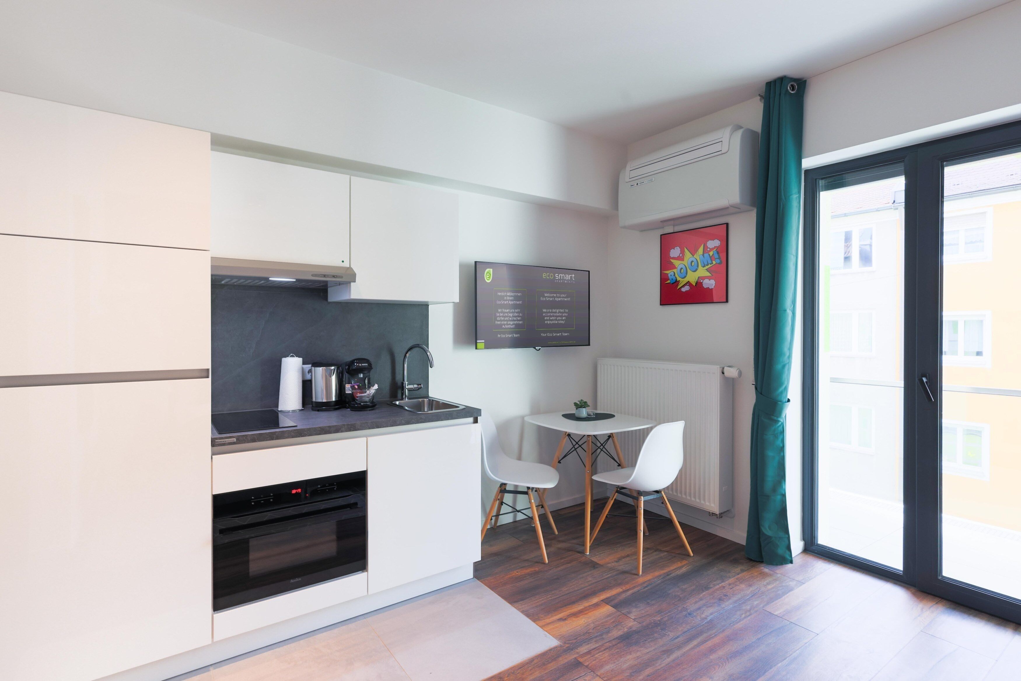 A modern kitchen with white cabinets, a built-in oven, and a coffee maker. There is a small dining table with two chairs and a window with a curtain.
