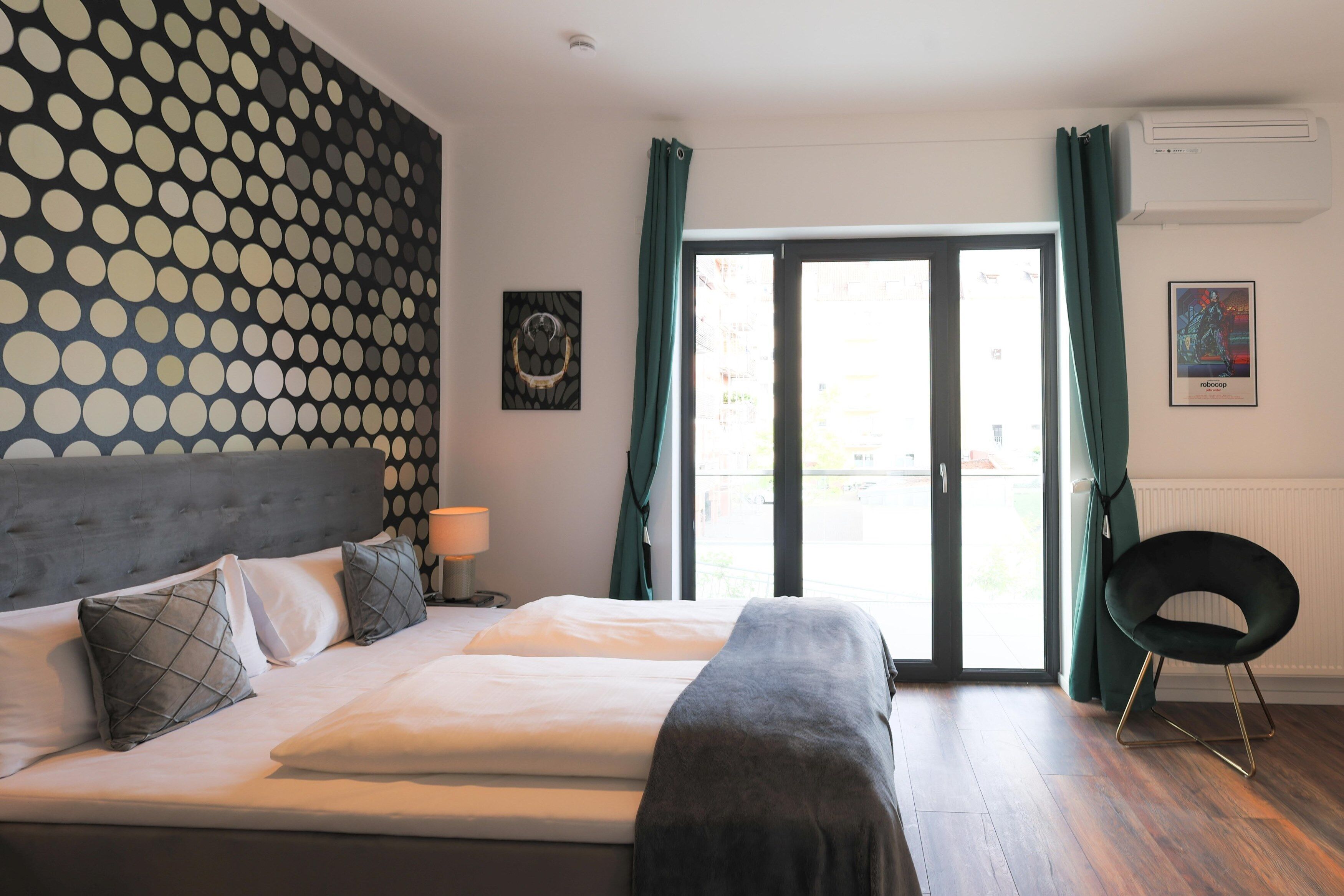 A bedroom with a bed, a chair, a window with curtains, and a wall with a polka dot pattern.