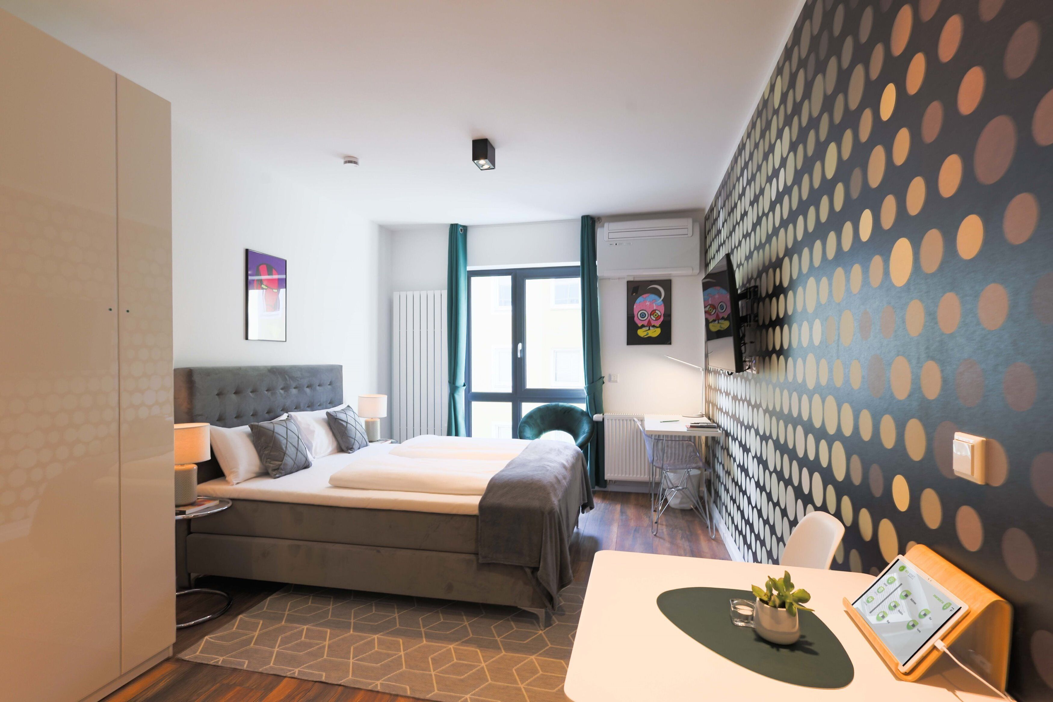 A modern hotel room with a large bed, a desk with a computer, a chair, and a small table with a plant.