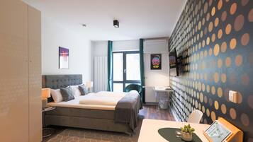 A modern hotel room with a large bed, a desk with a computer, a chair, and a small table with a plant.