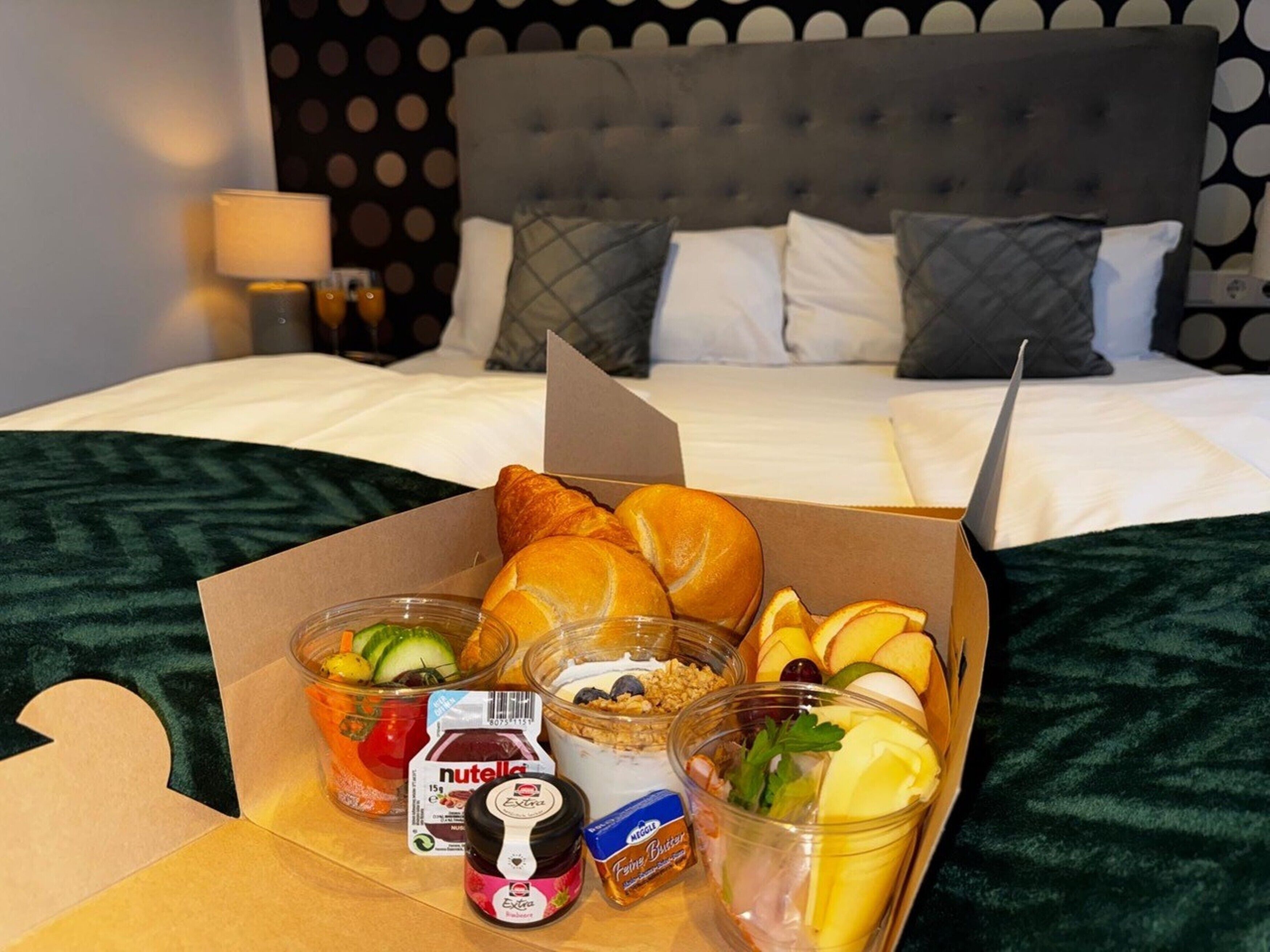 Une chambre d’hôtel équipée d’un lit, d’un ordinateur portable et d’une boîte contenant le petit-déjeuner, avec des croissants, des fruits, du yaourt et des confitures.
