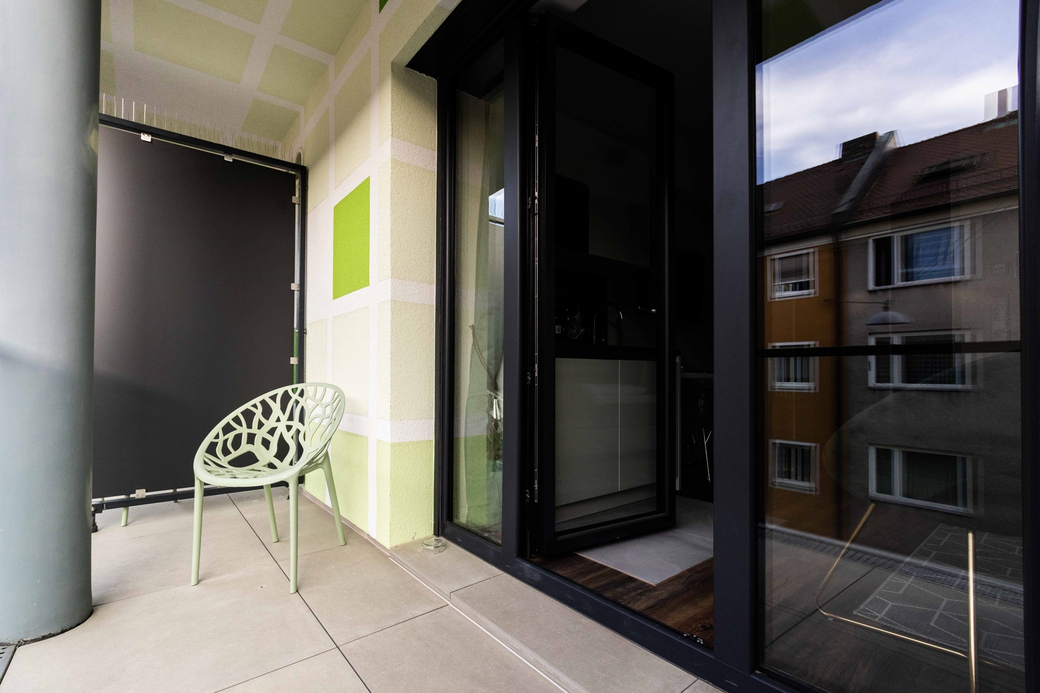 A modern apartment with a sliding glass door leading to a balcony, a green chair, and a view of neighboring buildings.