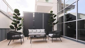 Un espace extérieur moderne, aménagé avec un canapé et deux fauteuils, une cloison en verre et des plantes ornementales en pot.