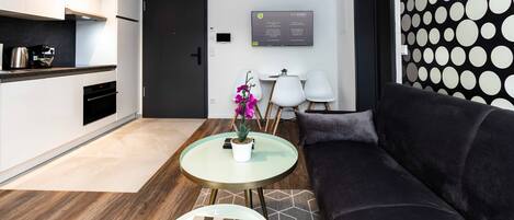 A modern living room with a dark sofa, a glass coffee table with a book, a small white table with chairs, and a kitchen area with white cabinets and a black door.