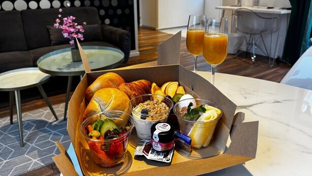 Une boîte contenant des croissants, des fruits, un yaourt et du jus, posée sur une table dans une chambre d’hôtel moderne, avec un canapé et un coin repas en arrière-plan.