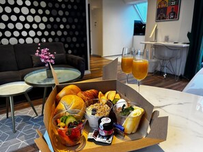 Una caja de desayuno con cruasanes, fruta, yogur y jugo, sobre una mesa en una habitación de hotel moderna con un sofá y un área de comedor al fondo.
