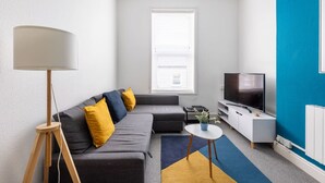 A modern living room with a grey sofa, yellow cushions, a floor lamp, a TV, and a small white table with a plant. - Judah Suite by Koya Homes (Barry)