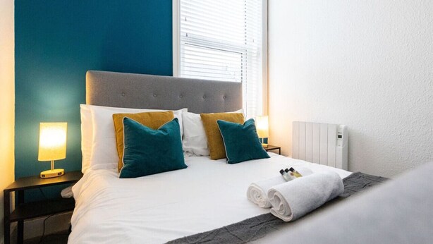 A neatly made bed with white linens and teal and mustard pillows, a bedside lamp, and a rolled towel on the bed. - Judah Suite by Koya Homes (Barry)
