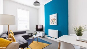 A modern living room with a sofa, a TV, a dining table, and a blue accent wall. - Judah Suite by Koya Homes (Barry)