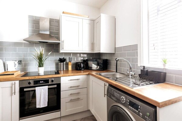 A modern kitchen with white cabinets, grey tiles, a built-in oven, a microwave, a dishwasher, and a window with blinds.