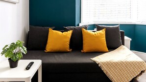 A dark-colored sofa with two yellow pillows, a white side table with a remote control and a potted plant, and a window with blinds.