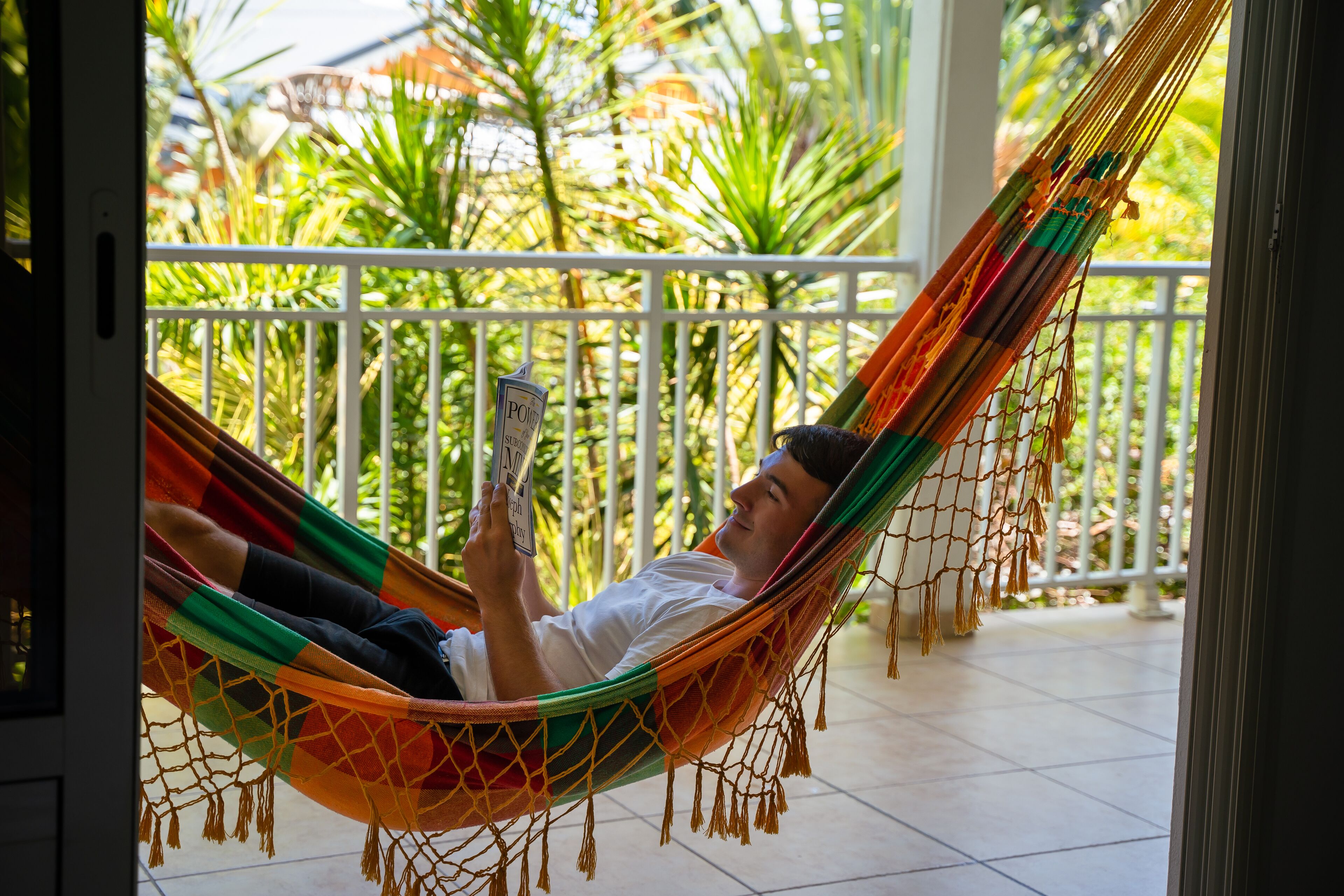 Una persona recostada en una hamaca en un balcón, con vista a plantas tropicales.