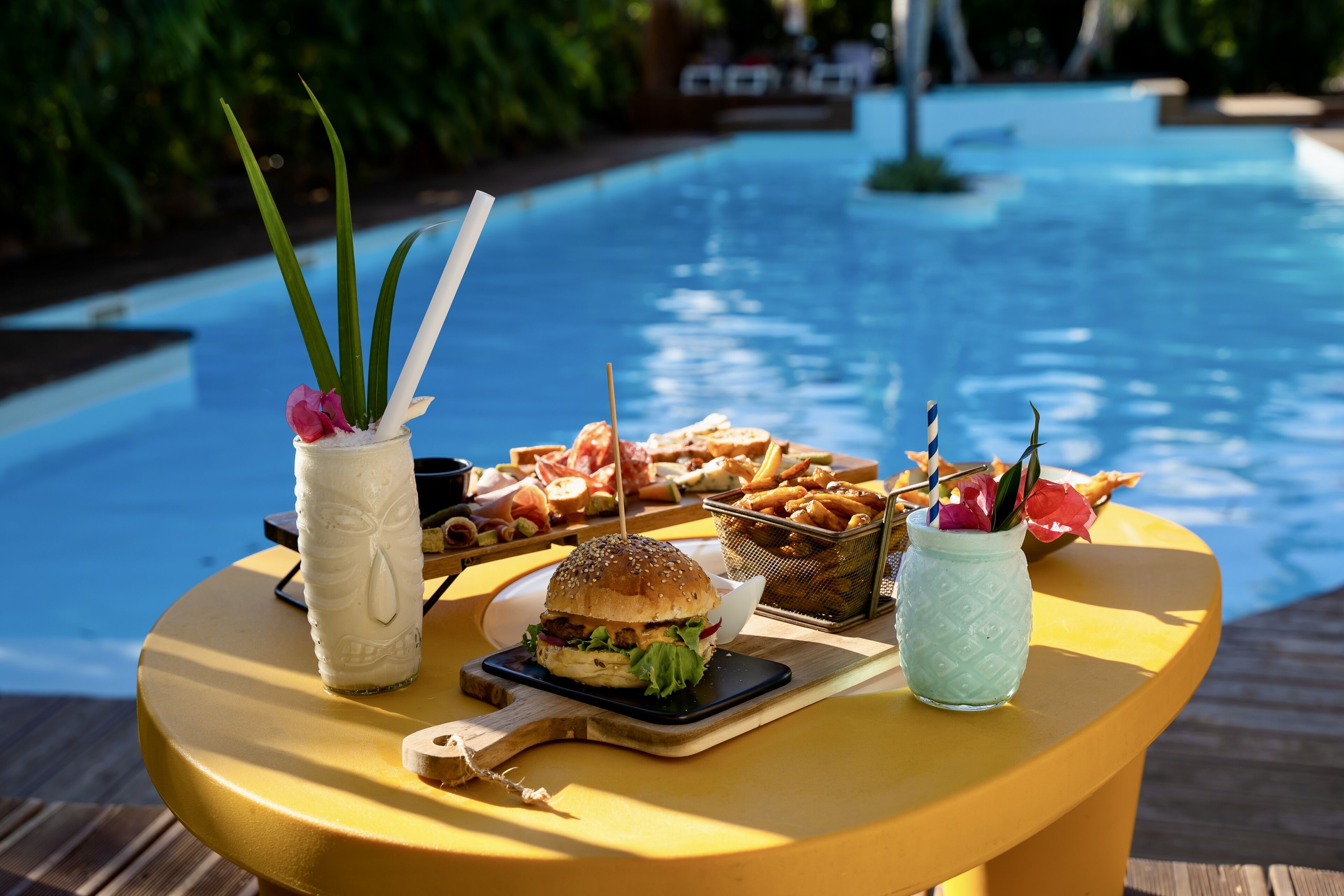 Une table à manger avec un hamburger, des frites et des boissons, placée au bord d’une piscine.