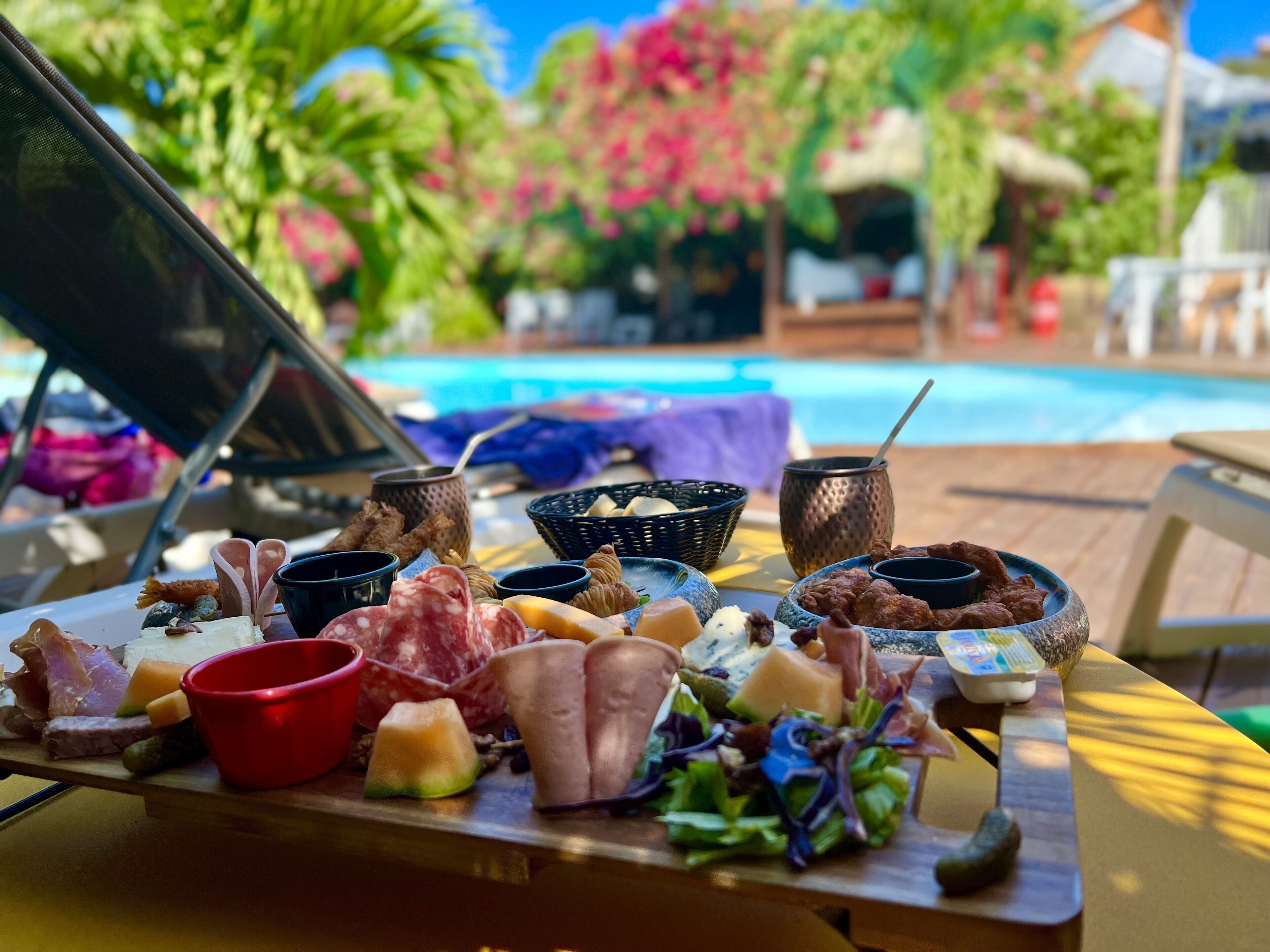 Una mesa de buffet con varios quesos, carnes y frutas, frente a una vista con piscina, palmeras y cielo despejado.