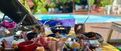 Una mesa de buffet con varios quesos, carnes y frutas, frente a una vista con piscina, palmeras y cielo despejado.
