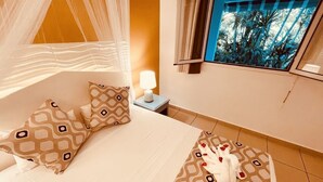 A hotel room with a bed, patterned pillows, a nightstand with a lamp, and a window with a view of palm trees. - Tropicana Suites (Deshaies)