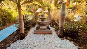 A garden with a decorative fountain, palm trees, and a pathway. - Tropicana Suites (Deshaies)